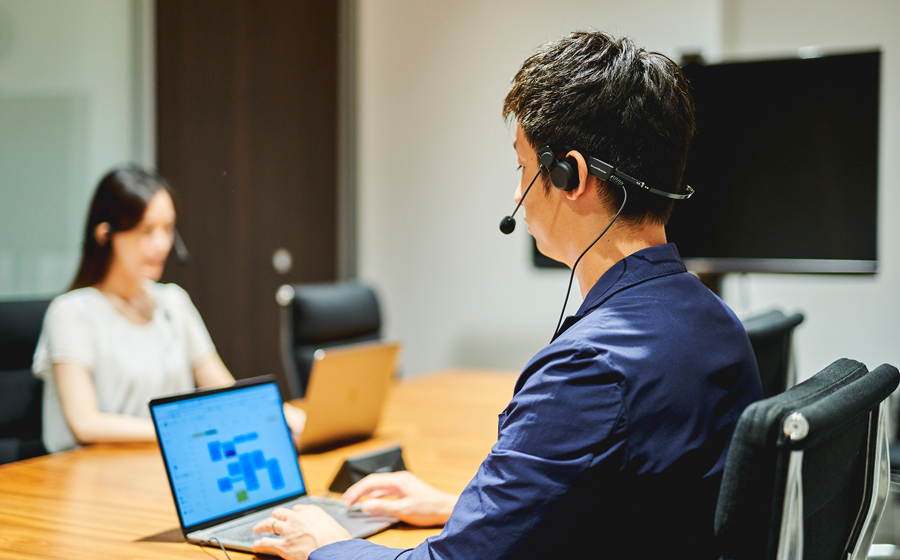 男性と女性がオフィスにいて、男性は城下工業SOUND WARRIORの折りたたみ式片耳日本製ヘッドセットSW-HW2をつけパソコンでスケジュール管理をしている