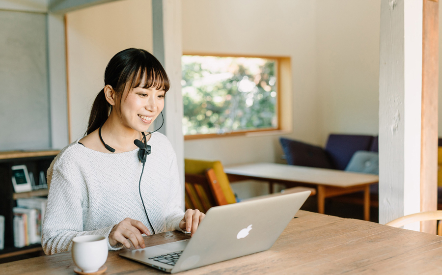 カフェの中で夏用のセーターを着た女性が日本製の城下工業 SOUND WARRIORの1ヘッドセットマイクSW-NS1を首にかけ、ノートパソコンに向かって微笑んでいる