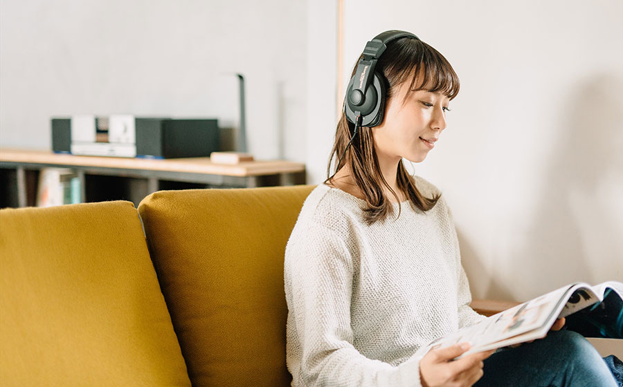 城下工業SOUND WARRIORモニターヘッドホンSW-HP20-Bを女性が装着しリビングで雑誌を読んでいる