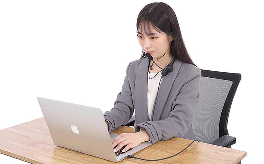 女性が日本製の城下工業 SOUND WARRIORのSW-NS1を首にかけ、両耳イヤホンをつけてノートパソコンで仕事をしています