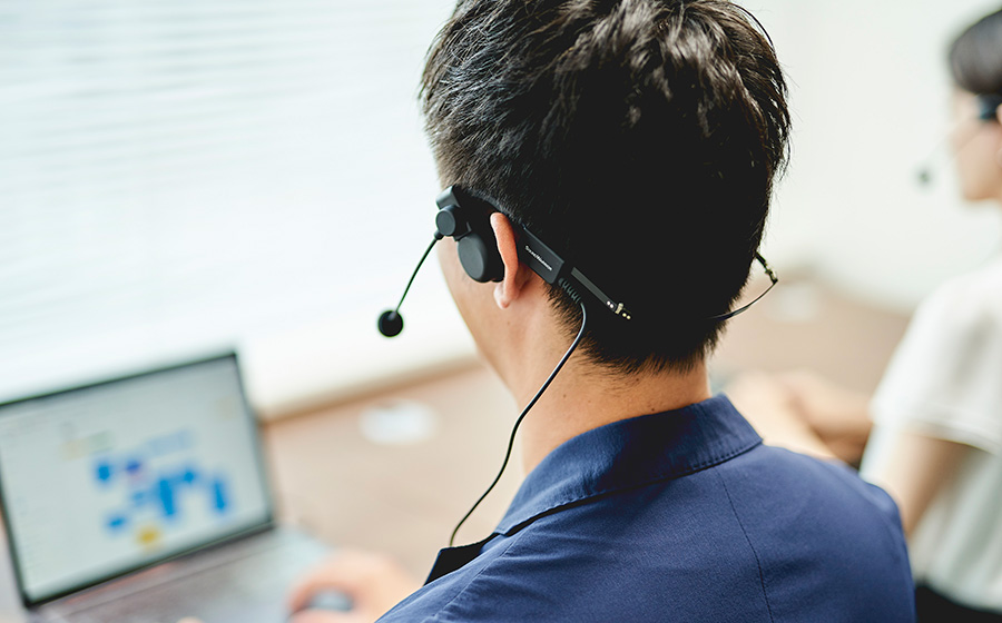 城下工業SOUND WARRIORヘッドセットは片耳のオンイヤーヘッドホンタイプで、男性がシェアオフィスでヘッドセットをつけてパソコンをしている